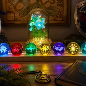 Può includere: Sette sfere luminose di diversi colori, ognuna con un design unico, poggiano su una base di legno. Una cupola di vetro con fiori e luci di fata è dietro le sfere. Sono visibili anche una spada, uno scudo e un libro.