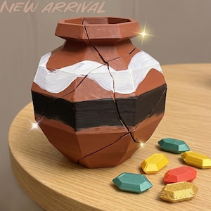 May include: A faceted terracotta-colored vase with a wide opening, featuring a horizontal white painted wavy stripe and a black painted band. The vase has visible structural seams and is accompanied by five small, gem-shaped plastic tokens.