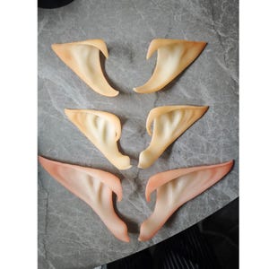 Puede incluir: Seis prótesis de orejas de elfo en varios tonos de piel, desde beige claro hasta rosa claro. Las orejas están detalladas con crestas internas y están dispuestas sobre una superficie gris.