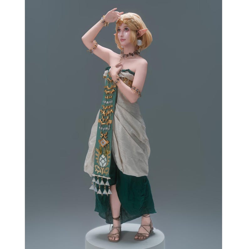 Op de afbeelding: Een blond elfenfiguur met puntige oren draagt een strapless, gelaagde outfit in beige en donkergroen. Het kledingstuk heeft een lang, verticaal groen paneel versierd met gouden geometrische patronen en bungelende driehoekige bedels.