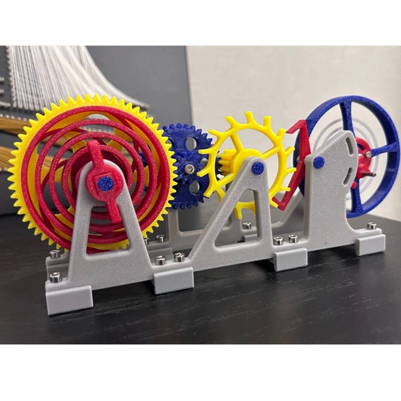 Op de afbeelding: Een kleurrijk 3D-geprint kinetisch tandwielmodel. Het model heeft een groot geel en rood tandwiel, een blauw tandwiel en een geel tandwiel, allemaal gemonteerd op een grijze basis. De tandwielen zijn ontworpen om in elkaar te grijpen en te draaien.