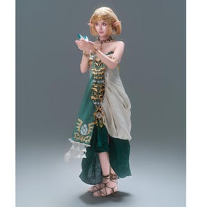 Op de afbeelding: Een kostuum in fantasystijl met een gelaagde jurk met een donkergroene rok, een cr&egrave;mekleurig lijfje en een decoratieve tabberd met gouden randen. De outfit bevat puntige oorprotheses, een gouden hoofdtooi en bruine sandalen met bandjes.