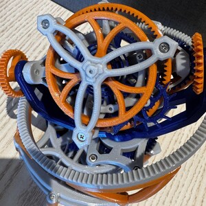 以下が含まれることがあります： オレンジ、青、グレーの色合いの、複雑で多層的な歯車機構。 複雑なデザインは、中央の3本スポークのホイールを含む、多数の連動する歯車を特徴としています。 歯車は小さなネジで固定されており、詳細な機械彫刻を作成しています。