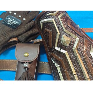 K&ouml;nnte beinhalten: Ein brauner, texturierter Armschutz mit metallisch silbernen geometrischen Akzenten, eine braune Ledertasche mit Messingdruckknopf und Quaste sowie ein brauner Strickhandschuh mit schwarzem Aufn&auml;her, wei&szlig;em Fl&uuml;gelwappen und silbernen Nieten.