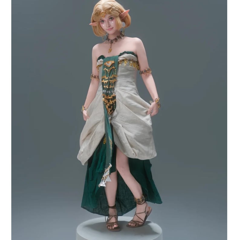 Op de afbeelding: Een beeldje van een blonde elf met puntige oren in een strapless jurk met een cr&egrave;mekleurig lijfje en een donkergroene rok. De jurk is voorzien van een centraal goudkleurig decoratief paneel, gouden armmanchetten, een ketting en bruine sandalen.