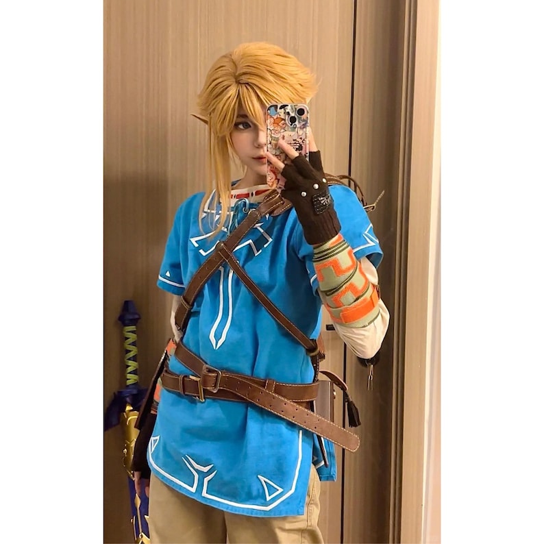 K&ouml;nnte beinhalten: Eine leuchtend blaue Tunika mit wei&szlig;em Saum, getragen mit einem braunen Ledergeschirr und G&uuml;rtel. Das Outfit umfasst einen braunen fingerlosen Handschuh, einen gemusterten &Auml;rmel und blondes Haar. Ein Schwertgriff mit blauen und goldenen Details ist sichtbar.