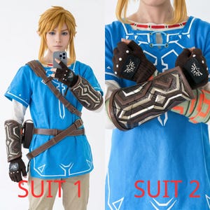 K&ouml;nnte beinhalten: Eine blaue Tunika mit wei&szlig;en geometrischen Mustern, braune fingerlose Handschuhe mit wei&szlig;en Nieten und einem schwarzen Emblem sowie ein brauner Unterarmschutz mit silbernen Metalldetails. Das Outfit umfasst ein braunes G&uuml;rtelsystem.