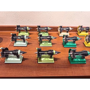 Puede incluir: Una colección de modelos en miniatura de motores a reacción. Cada modelo presenta un cuerpo negro con detalles plateados y verdes, montado sobre una base cuadrada. Las bases son de varios colores, incluyendo verde, amarillo y blanco. Los modelos están dispuestos sobre una superficie de madera.