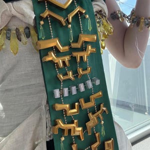 Op de afbeelding: Een donkergroene stoffen sjerp versierd met goudkleurige geometrische vormen, kleine doorzichtige rechthoekige blokjes en bungelende gele kristalachtige hangers. De sjerp ligt over een lichtgekleurd kledingstuk en een bijpassende armband is zichtbaar.