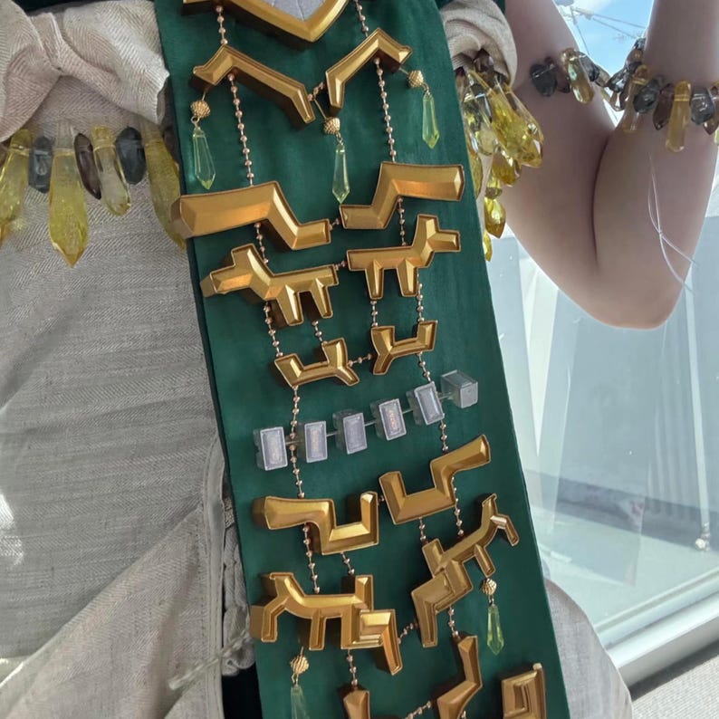 Op de afbeelding: Een donkergroene stoffen sjerp versierd met goudkleurige geometrische vormen, kleine doorzichtige rechthoekige blokjes en bungelende gele kristalachtige hangers. De sjerp ligt over een lichtgekleurd kledingstuk en een bijpassende armband is zichtbaar.