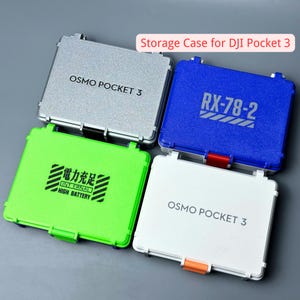 May include: Four storage cases for DJI Pocket 3 in silver, blue, green, and white. Each case has a rectangular shape with a hinged lid and a latch. The silver and white cases have "OSMO POCKET 3" printed on them. The blue case has "RX-78-2" printed on it. The green case has Chinese characters and "HIGH BATTERY" printed on it.