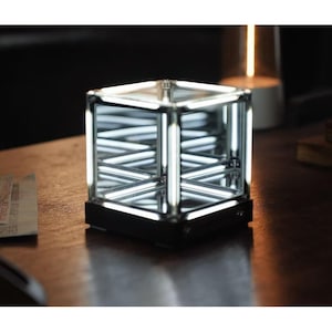 Peut inclure: Une lampe cube géométrique moderne avec une base noire et un intérieur en miroir. La lampe est dotée de lumières LED blanches vives disposées en croisillons, créant un effet d'infini. La lampe est posée sur une surface en bois.
