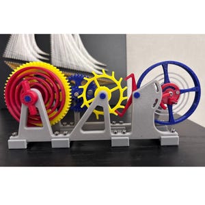 Może przedstawiać: Model mechaniczny wydrukowany w 3D, z szeregiem kolorowych kół zębatych i kołem balansowym. Koła zębate są żółte, niebieskie i czerwone, zamontowane na szarej podstawie. Koło balansowe jest niebiesko-czerwone. Model demonstruje zasady mechaniki.