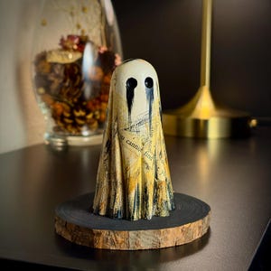 May include: A ghost-shaped figurine crafted from paper with printed text, featuring two hollow black eyes with black paint dripping downwards. The figure stands on a circular wooden base with a dark, charred-looking edge and natural bark.
