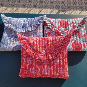 Può includere: Tre pochette in tessuto trapuntato con bordi a balze. Le pochette presentano motivi floreali in blu, rosso e rosa. Ogni pochette ha una chiusura con bottone. Le pochette sono disposte su una superficie verde scuro.