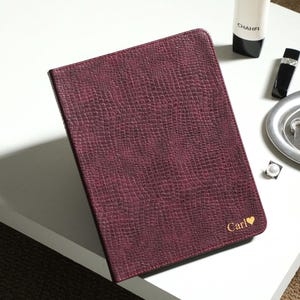 Pode incluir: Um caderno retangular de cor bordô profundo com uma capa texturizada com padrão de réptil. O canto inferior direito apresenta o texto dourado "Carl" seguido por um pequeno símbolo de coração. O caderno está posicionado em ângulo sobre uma superfície branca.
