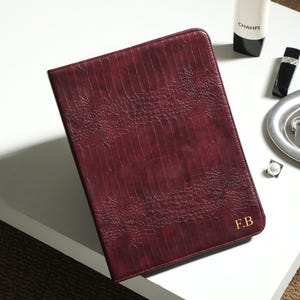 May include: A rectangular, dark burgundy portfolio case with a textured, crocodile-skin pattern. The front cover features the gold-embossed initials F.B. in the bottom right corner. The case has a structured, slim design with rounded corners.