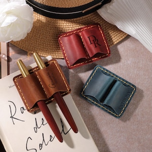May include: Three leather pen holders in brown, red, and blue, each holding pens. The brown holder rests on a book, while the others are on a neutral surface. The holders are personalized with initials and names.