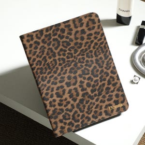 May include: A rectangular tablet or notebook case featuring a brown and black leopard print pattern. The bottom right corner of the case is personalized with the name Donna printed in gold lettering. The case has a smooth, flat surface.