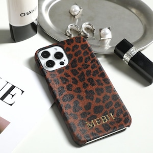 Peut inclure: Une coque de smartphone à motif léopard marron et noir avec une découpe pour triple appareil photo et le texte doré en relief MEBH en bas. La coque est placée à côté d'une page de magazine blanche, d'un plateau argenté avec des boucles d'oreilles.