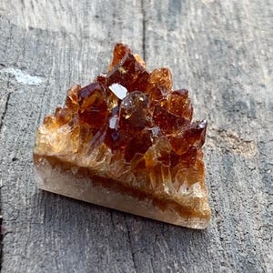 Op de afbeelding: Een driehoekige cluster van citrienkristallen, met amber- en goudbruine tinten. De kristallen hebben scherpe, puntige randen en een doorschijnende kwaliteit, rustend op een lichtgekleurde basis. De natuurlijke formatie staat op een verweerd houten oppervlak.