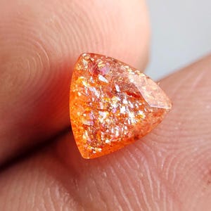 Può includere: Una gemma di pietra solare triangolare, di colore arancione con un aspetto scintillante e scintillante. La gemma lucidata è tenuta tra le dita. Il colore della pietra varia dall'arancione chiaro a un arancione rossastro più intenso.