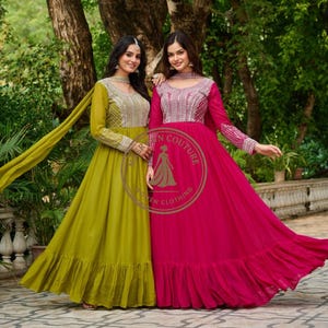 May include: Two women wearing long, flowing gowns. One gown is a vibrant fuchsia, the other a muted olive green. Both dresses feature intricate silver embroidery on the bodice and sleeves, with a ruffled hem. The image includes the text "Meerwan Couture".