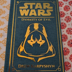 Pu&ograve; includere: Un libro con copertina rigida color verde acqua intitolato "Star Wars: Dynasty of Evil" con scritte e dettagli dorati. La copertina presenta un'illustrazione dorata dell'elmo di un personaggio. Il nome dell'autore, Drew Karpyshyn, &egrave; stampato in basso.