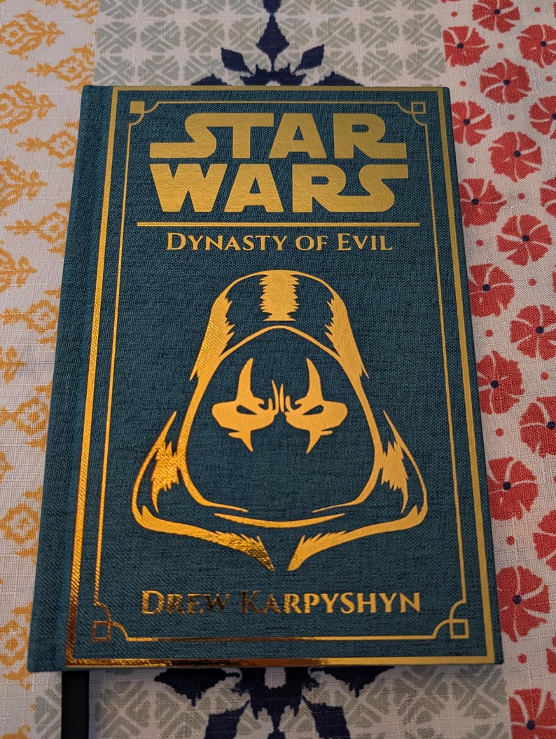 Pu&ograve; includere: Un libro con copertina rigida color verde acqua intitolato "Star Wars: Dynasty of Evil" con scritte e dettagli dorati. La copertina presenta un'illustrazione dorata dell'elmo di un personaggio. Il nome dell'autore, Drew Karpyshyn, &egrave; stampato in basso.