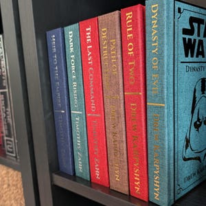 Pu&ograve; includere: Una collezione di libri di Star Wars su uno scaffale. I dorsi sono di vari colori, tra cui rosso, blu e marrone, con scritte dorate. I titoli includono "Dynasty of Evil" e "The Last Command".