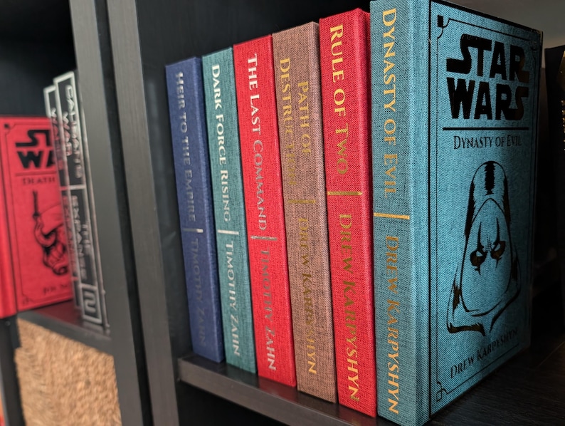 Pu&ograve; includere: Una collezione di libri di Star Wars su uno scaffale. I dorsi sono di vari colori, tra cui rosso, blu e marrone, con scritte dorate. I titoli includono "Dynasty of Evil" e "The Last Command".