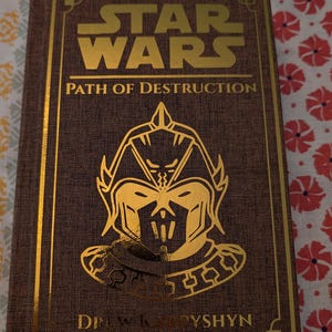 Pu&ograve; includere: Un libro con copertina rigida intitolato "Star Wars: Path of Destruction" con scritte e dettagli dorati. La copertina presenta un'illustrazione dorata di una figura con elmetto. Il nome dell'autore, Drew Karpyshyn, &egrave; stampato in basso.