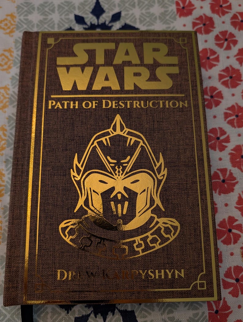 Pu&ograve; includere: Un libro con copertina rigida intitolato "Star Wars: Path of Destruction" con scritte e dettagli dorati. La copertina presenta un'illustrazione dorata di una figura con elmetto. Il nome dell'autore, Drew Karpyshyn, &egrave; stampato in basso.