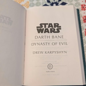 Pu&ograve; includere: Libro aperto con il logo di Star Wars e il titolo "Darth Bane: Dynasty of Evil" di Drew Karpyshyn. Sono visibili anche il logo Del Rey e "New York". Il libro ha una copertina verde acqua.