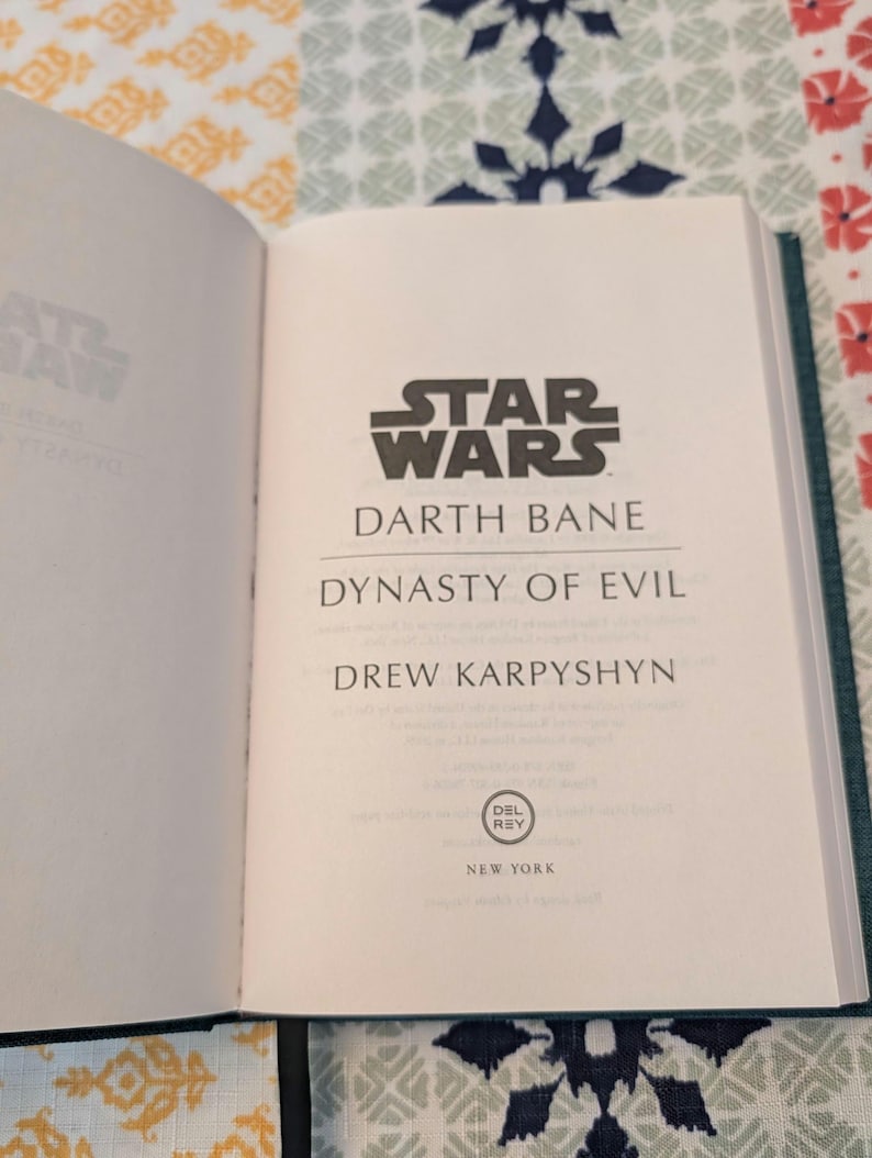 Pu&ograve; includere: Libro aperto con il logo di Star Wars e il titolo "Darth Bane: Dynasty of Evil" di Drew Karpyshyn. Sono visibili anche il logo Del Rey e "New York". Il libro ha una copertina verde acqua.