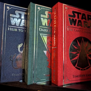 May include: A collection of four hardcover books featuring Star Wars titles including Ahsoka, Heir to the Empire, Dark Force Rising, and The Last Command. The books have textured covers in orange, blue, green, and red with metallic foil lettering.