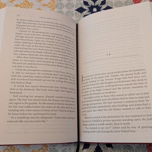 Pu&ograve; includere: Libro aperto con copertina rossa, che mostra testo su pagine color crema. Il libro &egrave; aperto su due pagine affiancate con testo, e i numeri di pagina sono visibili. Il libro &egrave; su una superficie a motivi.