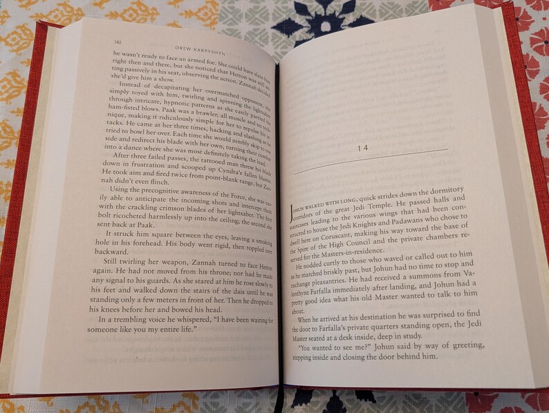 Pu&ograve; includere: Libro aperto con copertina rossa, che mostra testo su pagine color crema. Il libro &egrave; aperto su due pagine affiancate con testo, e i numeri di pagina sono visibili. Il libro &egrave; su una superficie a motivi.