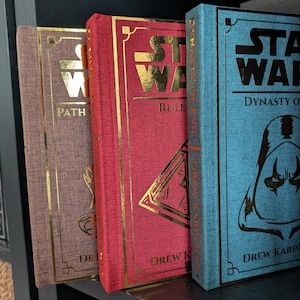 Pu&ograve; includere: Una collezione di libri con copertina rigida con scritte e dettagli dorati. I libri sono in vari colori, tra cui rosso, marrone e blu. I titoli includono "Star Wars" e "Dynasty of Evil". I libri sono esposti su una mensola scura.