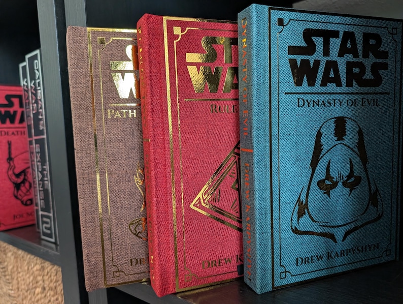 Pu&ograve; includere: Una collezione di libri con copertina rigida con scritte e dettagli dorati. I libri sono in vari colori, tra cui rosso, marrone e blu. I titoli includono "Star Wars" e "Dynasty of Evil". I libri sono esposti su una mensola scura.