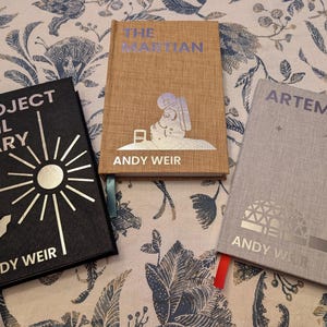 May include: Three hardcover books by Andy Weir are displayed on a floral patterned surface. The books are titled "Project Hail Mary", "The Martian", and "Artemis". Each book has silver lettering and unique cover art.
