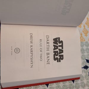 Pu&ograve; includere: Libro aperto intitolato "Star Wars: Darth Bane: Rule of Two" di Drew Karpyshyn. Il libro ha una copertina rossa e il logo di Star Wars &egrave; visibile. Il libro &egrave; aperto su una superficie a motivi.