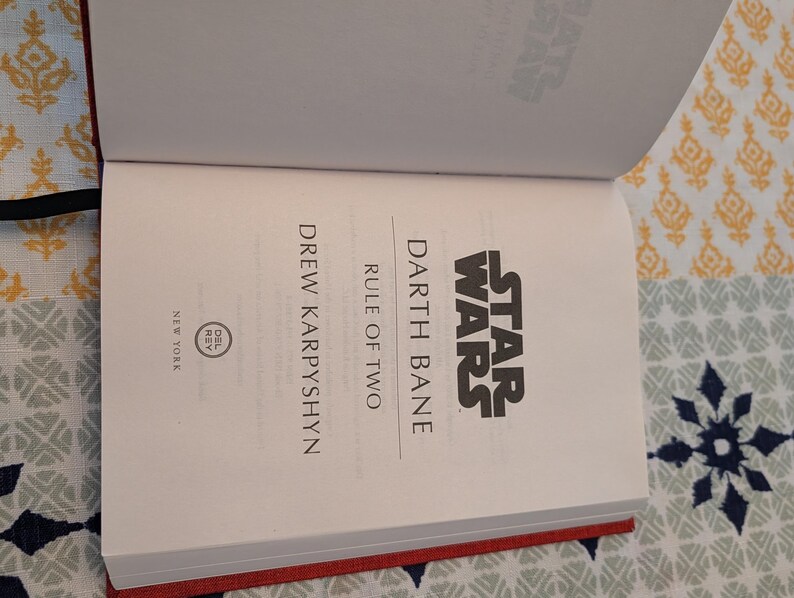 Pu&ograve; includere: Libro aperto intitolato "Star Wars: Darth Bane: Rule of Two" di Drew Karpyshyn. Il libro ha una copertina rossa e il logo di Star Wars &egrave; visibile. Il libro &egrave; aperto su una superficie a motivi.