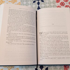 Pu&ograve; includere: Un libro aperto con testo sulle pagine, con una copertina turchese e un interno color crema. Il libro &egrave; aperto su una superficie a motivi. Il testo &egrave; in inglese, con il capitolo 14 visibile.