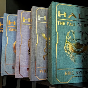 Peut inclure: Une rangée de cinq livres reliés Halo avec des couvertures en tissu texturé dans des tons bleu, violet et sarcelle. Chaque livre présente des lettres et des icônes graphiques en feuille d'or sur la couverture, dont le titre Halo: The Fall of Reach.
