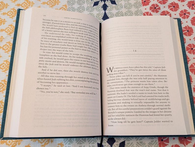 Pu&ograve; includere: Un libro aperto con copertina turchese e pagine color crema, appoggiato su una superficie a fantasia. Il testo &egrave; in inglese, con il capitolo 14 visibile. Le pagine del libro sono piene di testo.