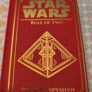 Pu&ograve; includere: Un libro con copertina rigida rossa con scritte dorate, intitolato "Star Wars Rule of Two". La copertina presenta il logo di Star Wars e un design geometrico dorato. Anche il nome dell'autore, Drew Karpyshyn, &egrave; in oro.