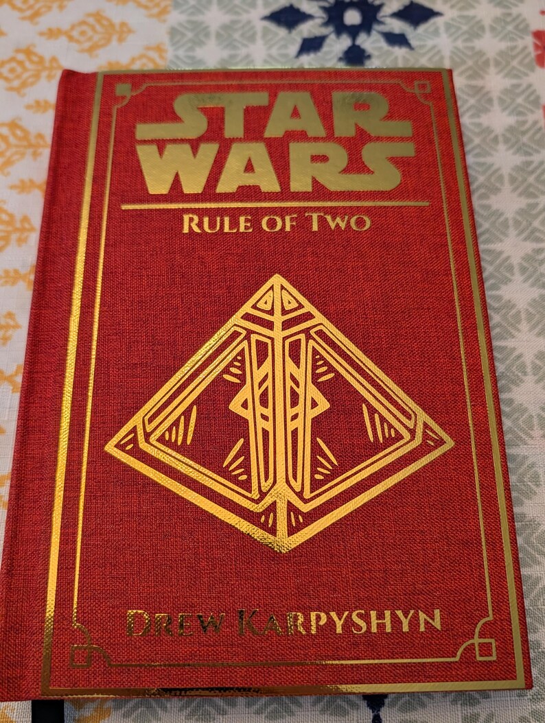 Pu&ograve; includere: Un libro con copertina rigida rossa con scritte dorate, intitolato "Star Wars Rule of Two". La copertina presenta il logo di Star Wars e un design geometrico dorato. Anche il nome dell'autore, Drew Karpyshyn, &egrave; in oro.