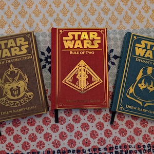 Pu&ograve; includere: Tre libri di Star Wars con copertina rigida con scritte e disegni dorati. I libri sono intitolati "Path of Destruction", "Rule of Two" e "Dynasty of Evil". Ogni libro ha una combinazione di colori diversa: marrone, rosso e verde acqua. Il nome dell'autore, Drew Karpyshyn, &egrave; stampato.