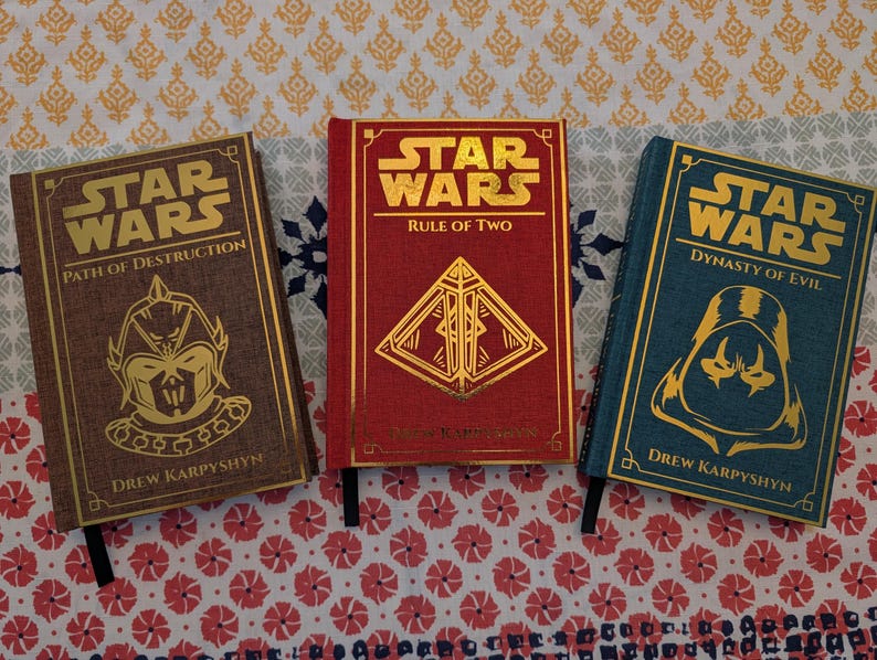 Pu&ograve; includere: Tre libri di Star Wars con copertina rigida con scritte e disegni dorati. I libri sono intitolati "Path of Destruction", "Rule of Two" e "Dynasty of Evil". Ogni libro ha una combinazione di colori diversa: marrone, rosso e verde acqua. Il nome dell'autore, Drew Karpyshyn, &egrave; stampato.